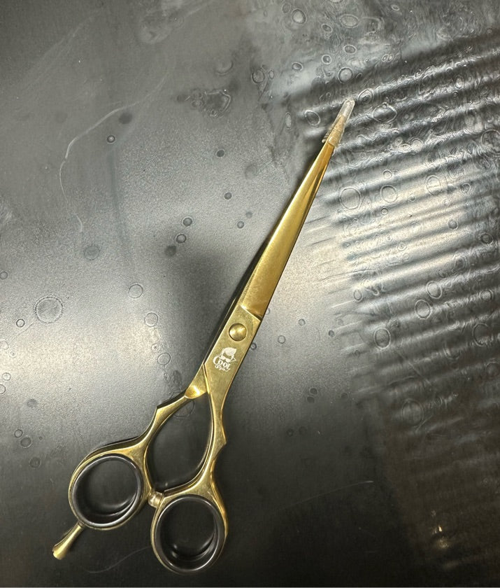 A pair of professional gold-handled scissors with sharp blades, resting on a textured surface.